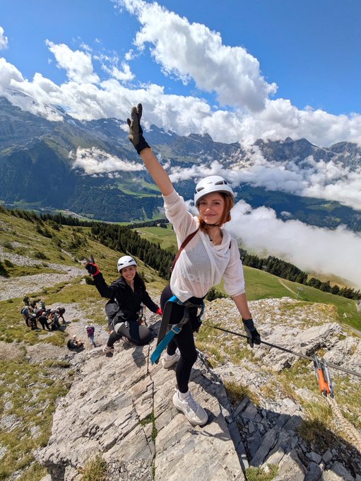 A woman in a white top and helmet waves with one arm raised while rock climbing on a mountain peak, with another climber behind her and a scenic alpine valley below.