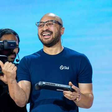 A bald man with glasses and a beard smiles while wearing a navy blue "Sui" t-shirt and holding a controller.
