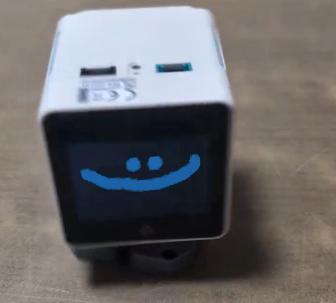 A small white cube-shaped electronic device with USB ports displaying a blue pixelated smiley face on its screen.