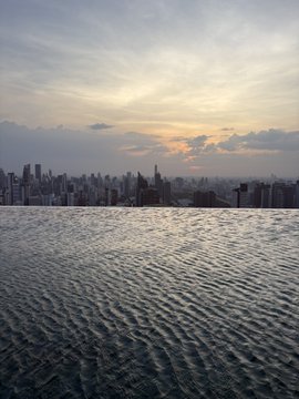 A view from an infinity pool looking out over a city skyline at sunset, with calm rippling water in the foreground and a hazy golden sky above.