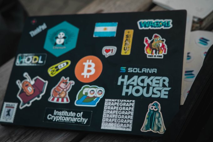 A black laptop covered in various cryptocurrency-themed stickers, including Bitcoin, Solana, HODL, WAGMI, and 'Solana Hacker House', with a 'Institute of Cryptoanarchy' sticker visible.