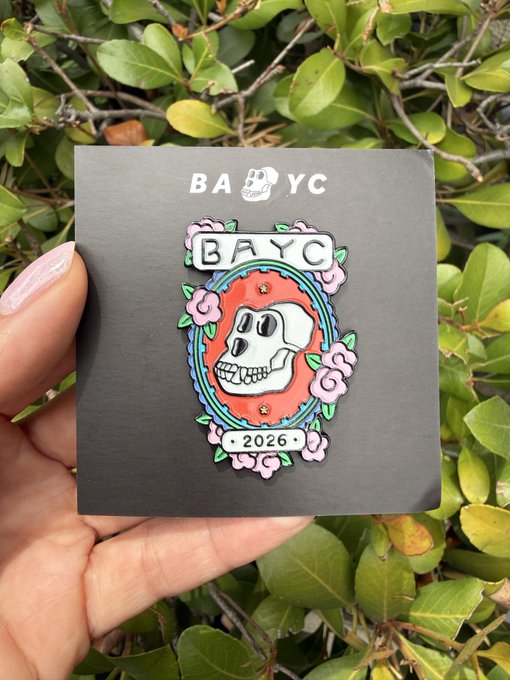 A hand holds a BAYC (Bored Ape Yacht Club) enamel pin featuring a skull and the text "BAYC" and "2026" against a backdrop of green foliage.