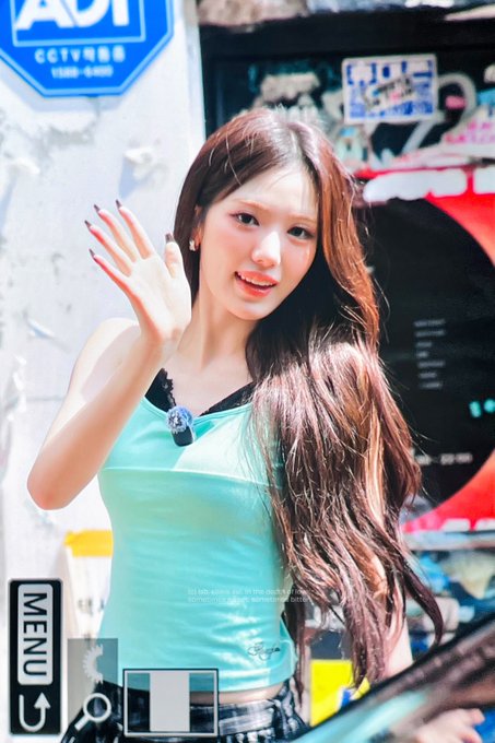 A young woman with long brown hair in a light blue top waving at the camera, with a CCTV sign visible in the background.