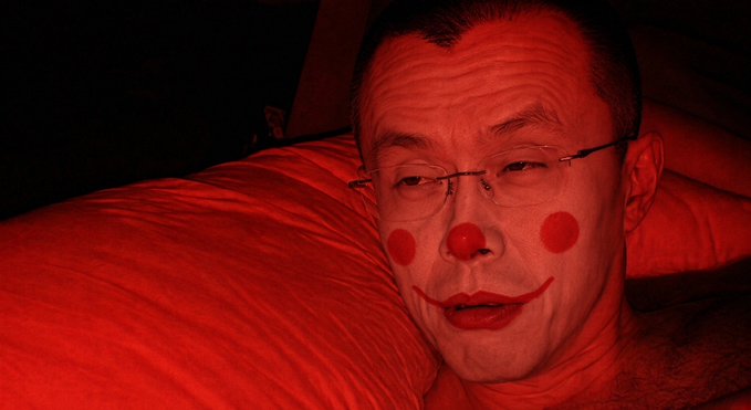 A man wearing red clown makeup, including a red nose, red lips, and red cheek circles, stares seriously at the viewer while resting his cheek against a red pillow, with the image bathed entirely in red lighting. The caption "CZ strikes again" likely references Changpeng Zhao, former head of the cryptocurrency platform, humorously reimagined in this eerie red-toned clown persona.