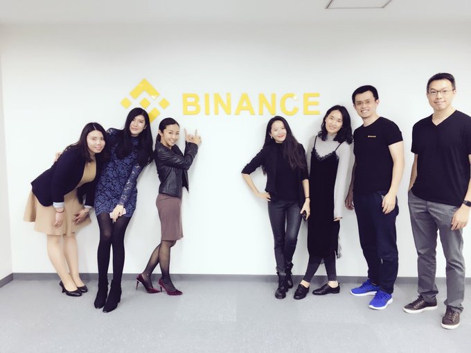 Seven people pose playfully in front of a yellow Binance logo on a white office wall, smiling and making gestures toward the camera. The accompanying caption references the early days of the Binance Charity partnership and a collaborative book project with proceeds donated to charity.
