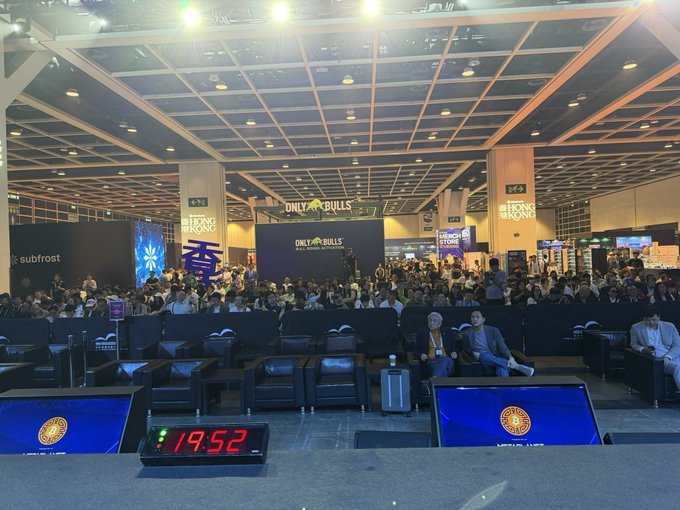A packed audience sits in a conference hall at the Bitcoin Asia 2025 event, with a "ONLY BULLS" stage display and a countdown clock showing 19:52 in the foreground, while sponsors like Hong Kong and subfrost are visible on banners around the venue.