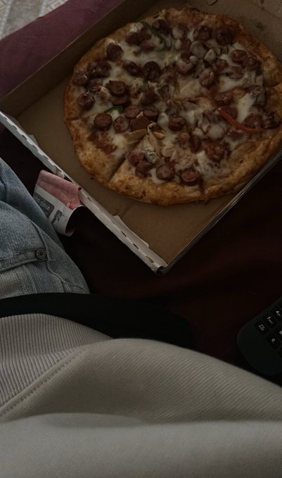An open pizza box on someone's lap containing a partially eaten pizza topped with sausage, cheese, and peppers, with a remote control visible to the right.