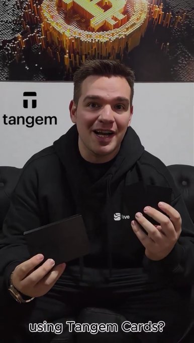 A man wearing a black hoodie with the Tangem logo sits in front of a Tangem-branded backdrop displaying a Bitcoin graphic, holding two black cards while a video caption asks 'using Tangem Cards?'.
