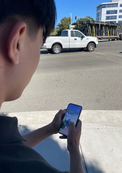 A young person with short dark hair stands on a sidewalk, playing Pokémon GO on their smartphone while a white pickup truck and a flatbed truck drive by on the street.