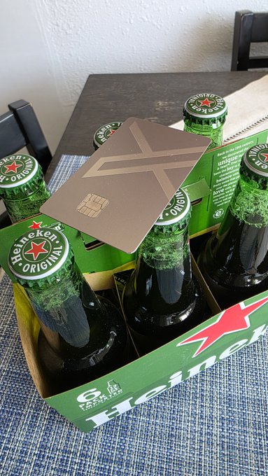 A six-pack of Heineken Original beer bottles in a green cardboard carrier on a table, with a metallic credit card-shaped object resting on top of the bottles.