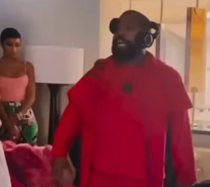 The image shows a man wearing red clothing, including a hoodie and sweatpants, standing in a living room with headphones around his neck. A woman can be seen behind him, possibly watching or waiting for her turn to perform. The scene appears to be set up for a family dinner, as indicated by the presence of a dining table and chairs nearby.
