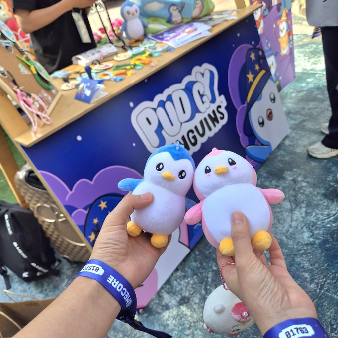 Two hands hold up small Pui Pui Penguins plush toys (one blue, one pink) at what appears to be a merchandise booth featuring Pui Pui Penguins branding in the background.