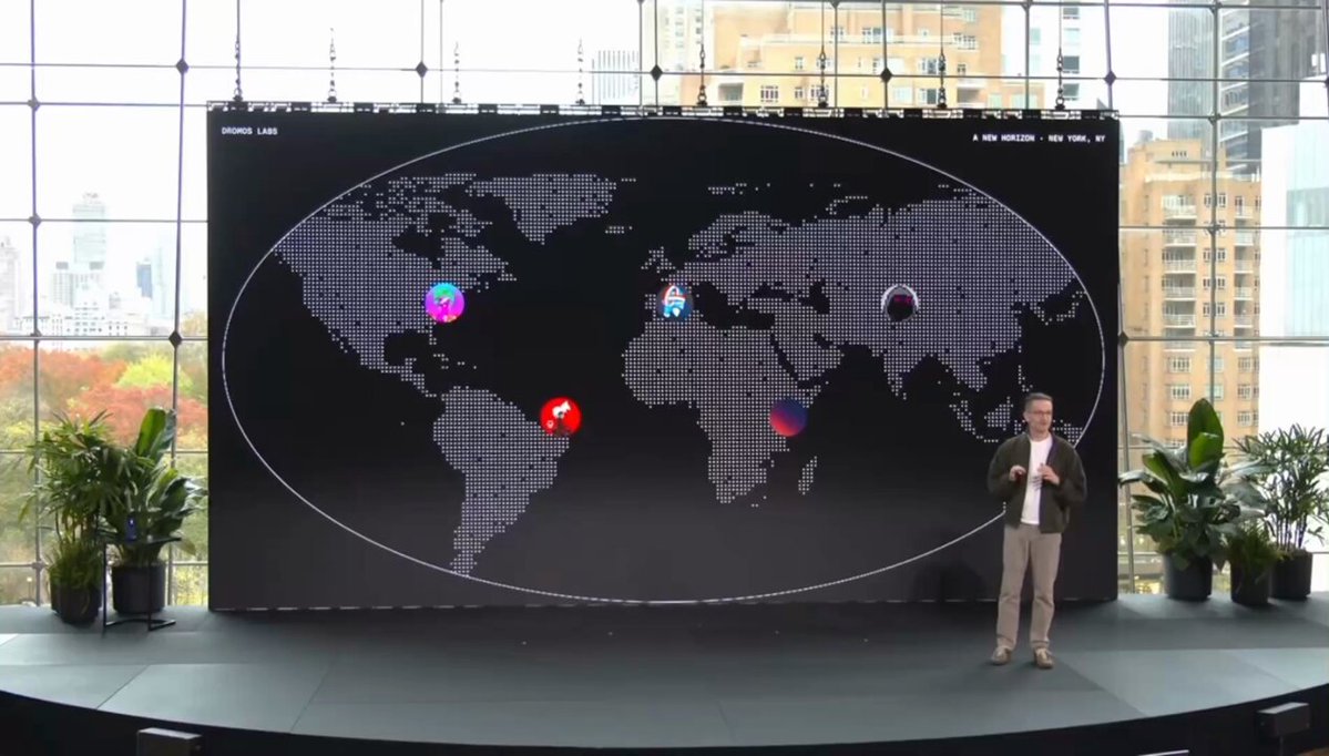 The image shows a man standing next to a large screen displaying a map of the world with various countries and cities marked. The man appears to be giving a presentation or speech about the importance of decentralized open networks, such as Ethereum, which can create a more inclusive and accessible online environment for people from different backgrounds and locations.