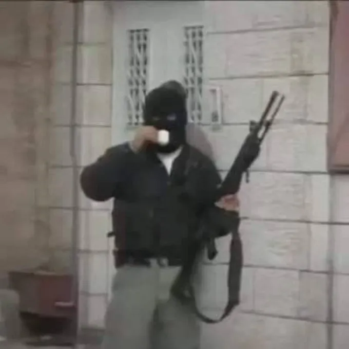 A grainy photo of an armed individual wearing a black balaclava, drinking from a white cup while holding a rifle, standing in front of a building with barred windows.