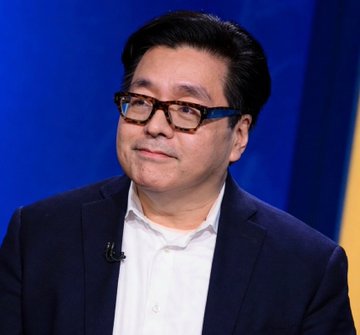 A man with black hair and black-framed glasses wears a navy blue suit jacket over a white shirt, photographed against a blue background.