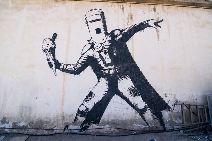 A black stencil graffiti art on a weathered wall depicting a figure in a welding helmet holding an ice cream cone while striking a dynamic pose.