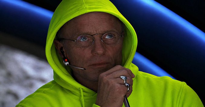 A man with a serious, contemplative expression wears a bright neon yellow/green hoodie and wire-frame glasses, with a small earpiece microphone visible on his cheek. He rests his chin on his hand adorned with silver rings, appearing to be deep in thought—matching the post's joke about "Hoodilio" attempting to revive the NFT market at its lowest point.