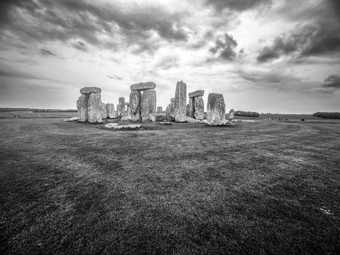 A dramatic black-and-white photograph of Stonehenge standing on a grassy plain beneath a moody, cloud-filled sky.