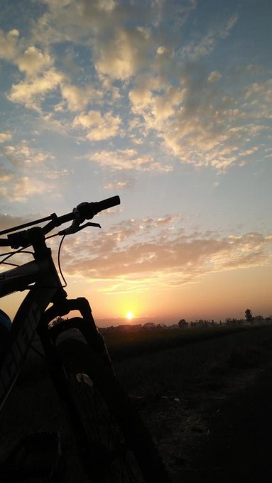 The image features a bicycle with its handlebars pointing towards the sky, which has a beautiful sunset behind it. Below the bike, there are some clouds that add depth to the scene. The photo appears to be taken during the day, as indicated by the presence of sunlight and shadows. The post seems to be promoting cycling or sharing an interesting moment captured in time with the sunset.