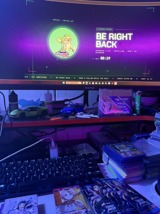 A computer monitor displays a Twitch stream overlay reading 'BE RIGHT BACK' with a countdown timer, alongside a cartoon character, sitting above a desk with a keyboard, toy cars, and anime merchandise including 'Jujutsu Kaisen' and 'Undead Unluck' items.