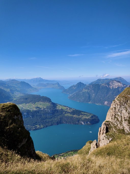 A scenic mountain overlook showing a deep blue lake winding through green valleys under a clear sky.