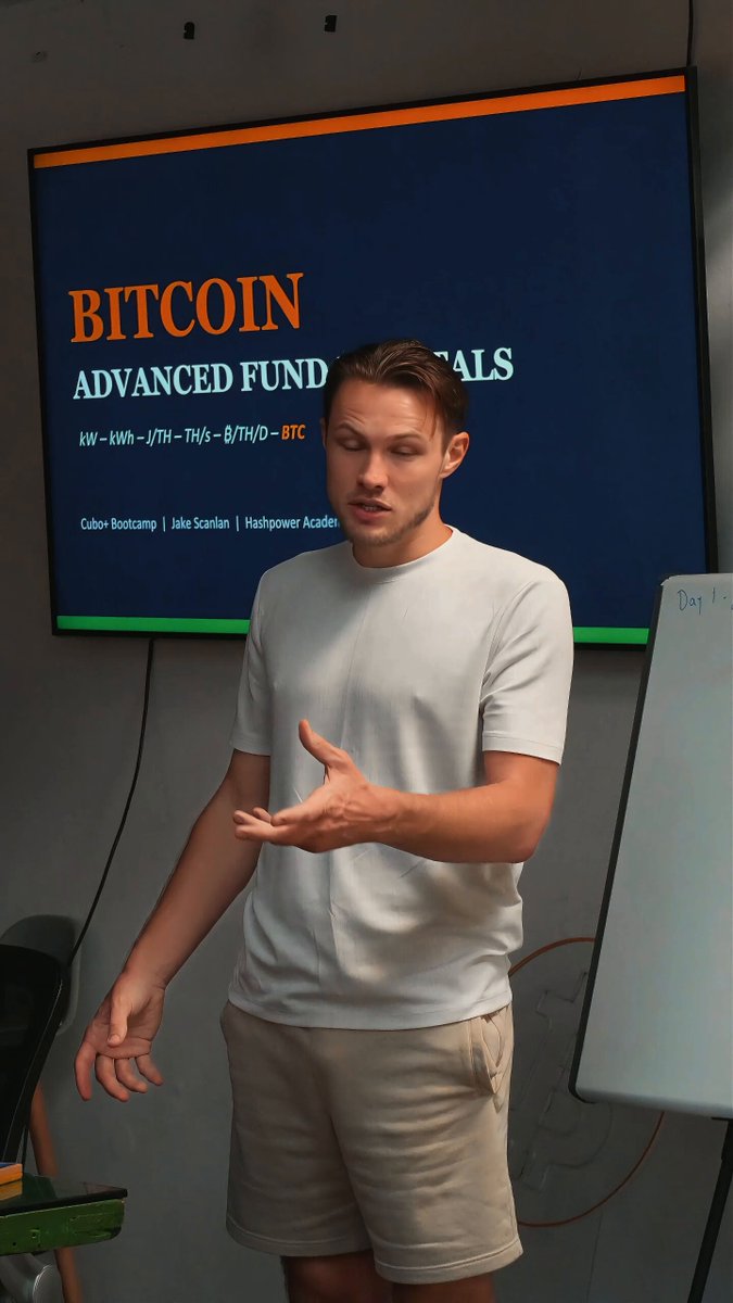 The image shows a man standing in front of a computer screen with a presentation about Bitcoin. The man appears to be giving a speech or lecture, possibly at an event like Bitcoin Week. He is wearing a white shirt and khaki shorts. The background features a large monitor displaying information related to Bitcoin, such as the words "BITCOIN" and "ADVANCED FUNDS".