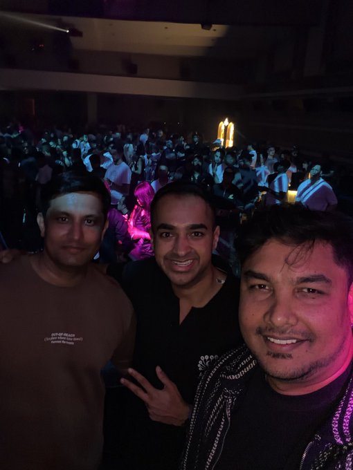 Three smiling friends take a selfie at the lively Token2049 conference during Token Week, with a packed auditorium and neon-lit stage visible behind them, capturing the energetic atmosphere of the event's first official day.