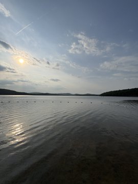 A serene lake scene at sunset with the sun low in the sky, casting a reflection on the calm water with hills in the distance.