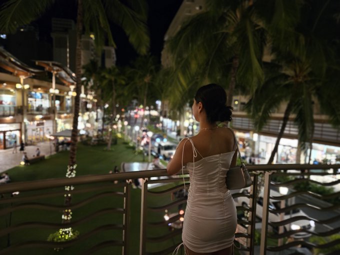A woman in a white dress stands at a balcony overlooking a tropical street at night, surrounded by palm trees and illuminated storefronts.