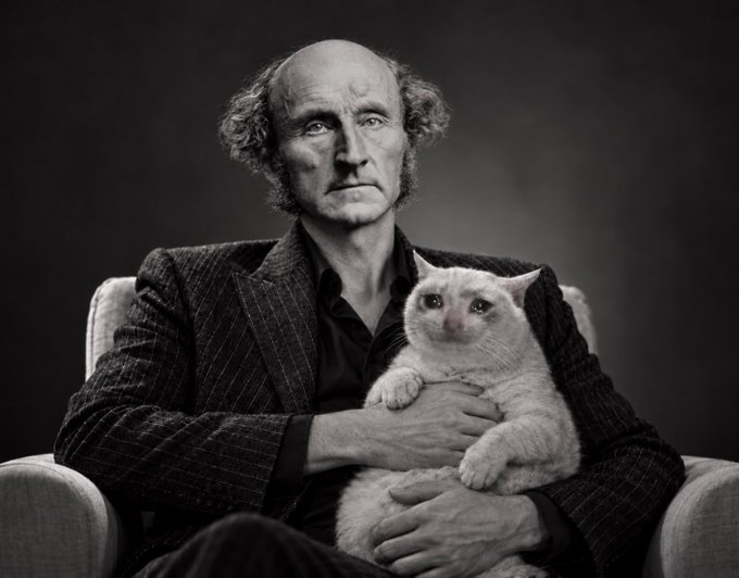 A black-and-white portrait shows a balding man in a pinstripe jacket sitting in a chair while holding a fluffy light-colored cat in his arms.