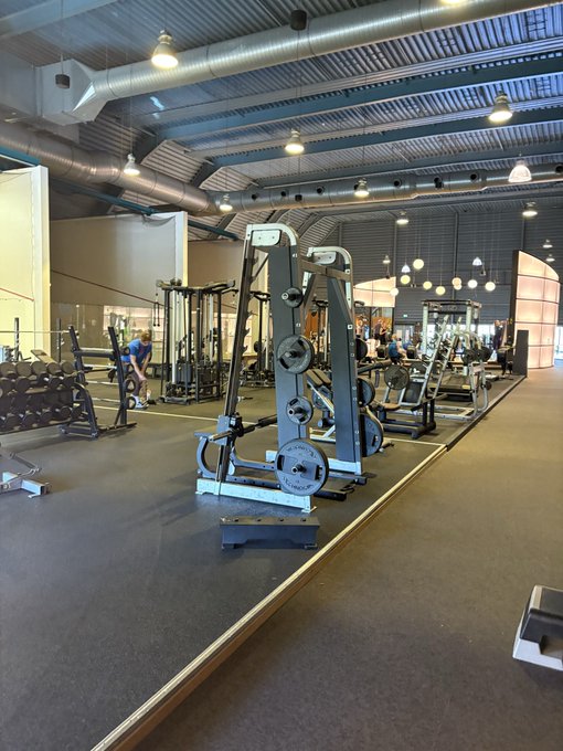 An interior view of a modern gym with various weight machines, mirrors, and industrial ceiling fixtures.