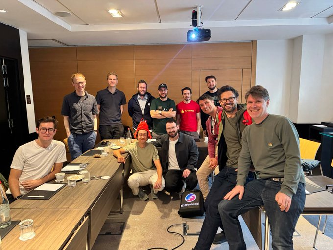 The image shows a group of men gathered around a conference table with a projector screen behind them. They are posing for a photo together, likely after attending an event or meeting related to cryptocurrency and blockchain technology. The room has a wooden ceiling and walls, giving it a warm and inviting atmosphere. In the background, there is a chair visible on one side of the image.