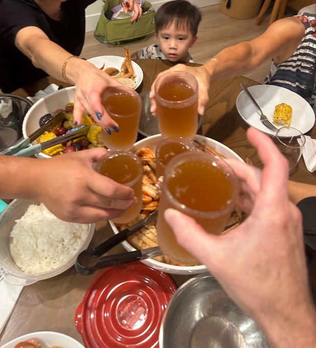 A group of people clink small glasses filled with an amber beverage in a toast over a dinner table laden with corn on the cob, rice, and other dishes, while a young child watches from the background.
