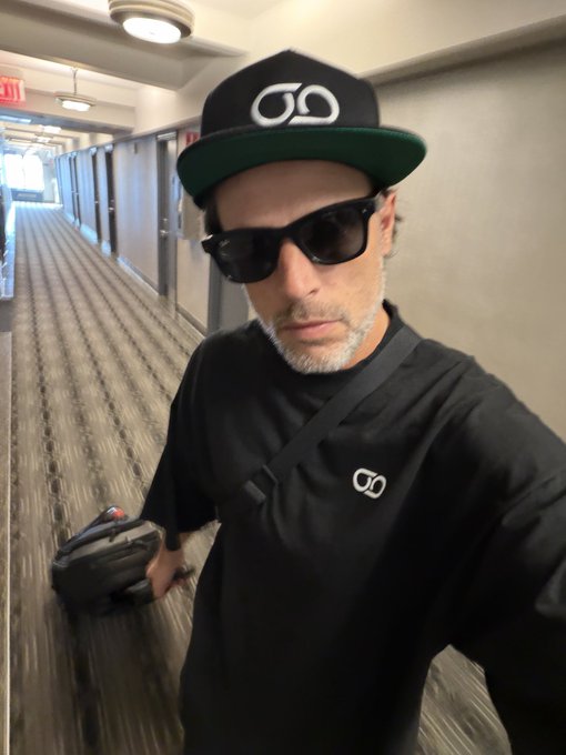 A man takes a selfie in a hotel hallway wearing a black t-shirt, sunglasses, and a black and green cap, both featuring a matching eyeglasses-style logo.