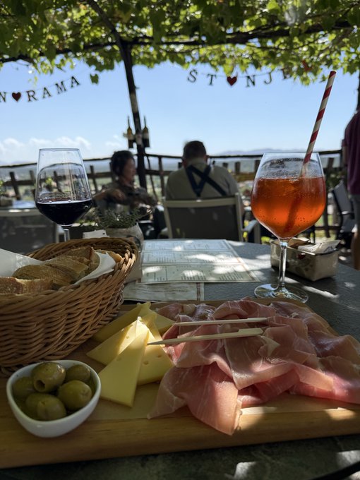 An outdoor dining scene featuring a wooden board with prosciutto, cheese, and olives, alongside a glass of red wine and an orange cocktail with a striped straw, with 'STONJA' banners visible overhead.