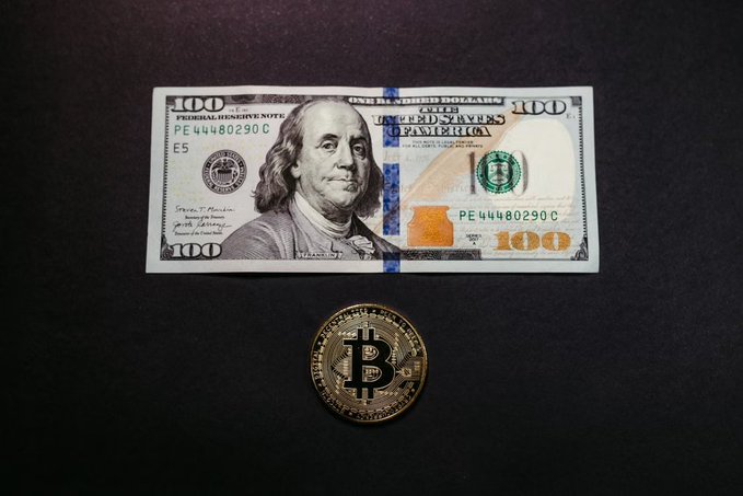 A $100 US bill placed above a physical Bitcoin coin against a black background.