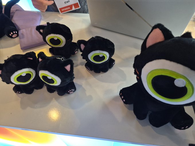 Several black plush toys with large green eyes, resembling cartoon cats, sit on a table at what appears to be a convention booth, with one held in the foreground.