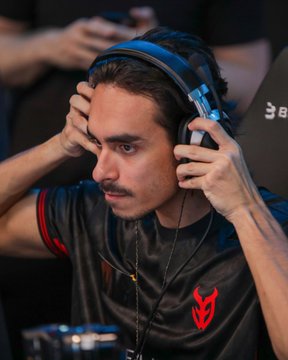 A male esports player with a mustache wearing a black jersey and blue headset adjusts his headphones with a focused expression during a match.