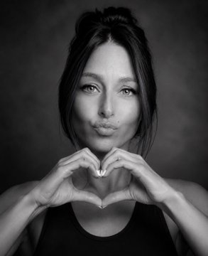 A black and white portrait photograph of a woman with dark hair pursing her lips and forming a heart shape with her hands.