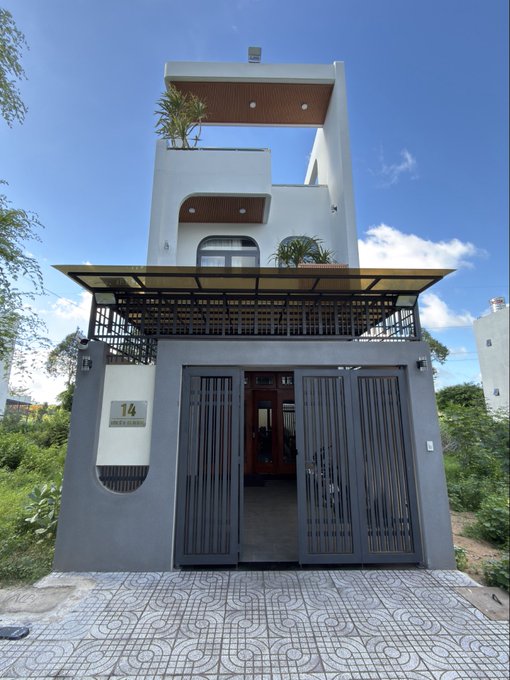 A modern three-story white house with dark gray accents, numbered 14, featuring a metal gate entrance and a balcony with plants.