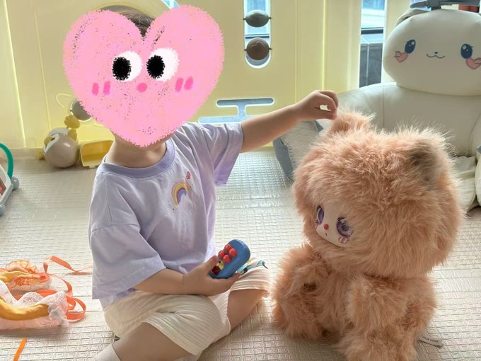 A child with a pink heart sticker covering their face plays with a small toy next to a fluffy brown teddy bear, seated on a play mat with a white plush toy in the background.