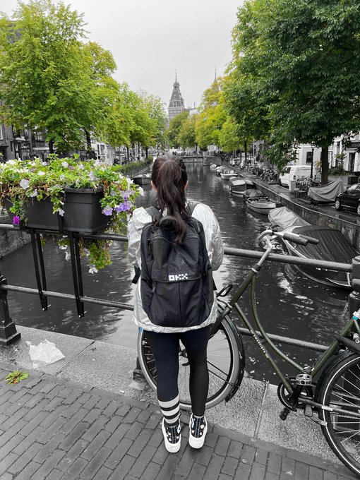 A woman with a black "DKXX" backpack stands on a bridge overlooking a picturesque Amsterdam canal, ready to give the bag away in a Europe-themed giveaway reaching for 10K likes.