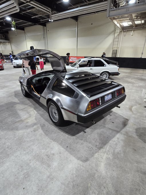 A silver DeLorean DMC-12 sports car with its gull-wing doors open is displayed at what appears to be a car show inside a large warehouse.