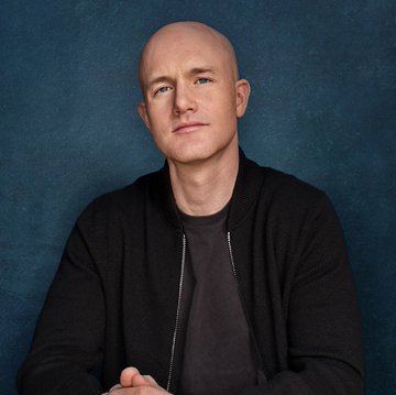 A professional headshot of a bald man in a dark zip-up jacket against a blue textured background, likely representing a company executive in relation to the breaking news about Coinbase's massive $52 million Bitcoin purchase, bringing their holdings to 17,311 BTC.