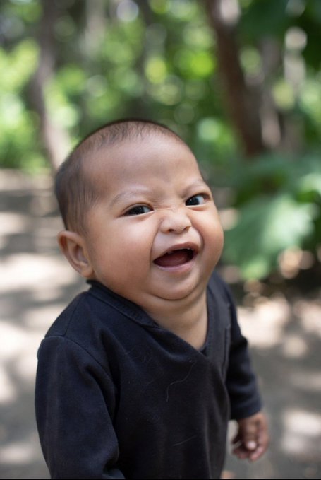 A close-up portrait of a smiling, dark-haired baby wearing a black shirt, set against a blurred green outdoor background.