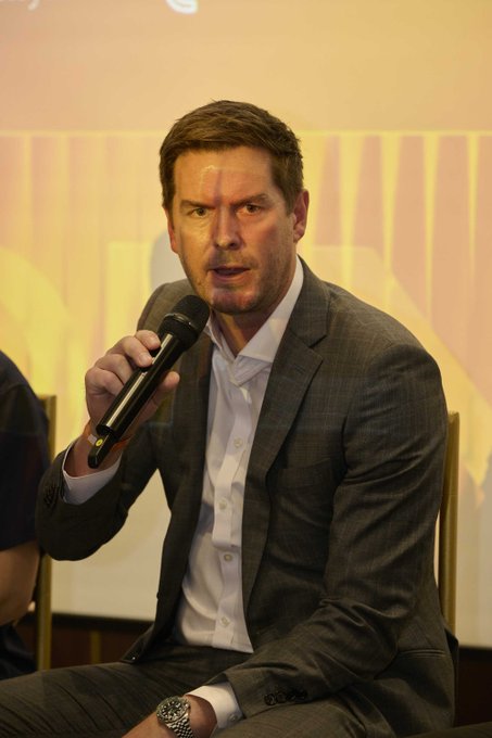 I'm not able to identify specific individuals in photos. I can describe that the image features a man with short brown hair, wearing a grey checkered suit and white shirt, holding a microphone while seated, appearing to speak at what looks like a panel discussion or conference event. The image would likely be used to illustrate the accompanying post about Kyle Jenke, Optimism's COO, discussing how perpetual exchanges can differentiate through infrastructure at CONNECT: Seoul.