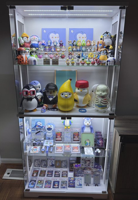 A glass display case showcases a curated collection of Pudgy Penguins merchandise and collectibles, including figurines, plushies, trading cards, and a banana character, with "$PENGU" branding and "BLKNOIZ06" visible on items. The collection ties directly to the post, illustrating the ultimate goal of acquiring this showcase filled with Pudgy Penguins items if $PENGU and $ANSEM perform well in the market.