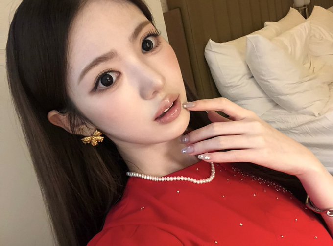 A young woman with long dark hair wearing a red top and gold bee earrings takes a close-up selfie.