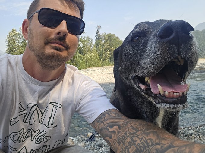A selfie of a man wearing sunglasses and a white t-shirt with tattooed arms, posing next to a happy black Labrador by a river.