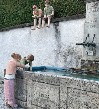 Two sculptures of children playing at a stone fountain dated '1924', with two children sitting on the wall above and two leaning over the basin filled with water.